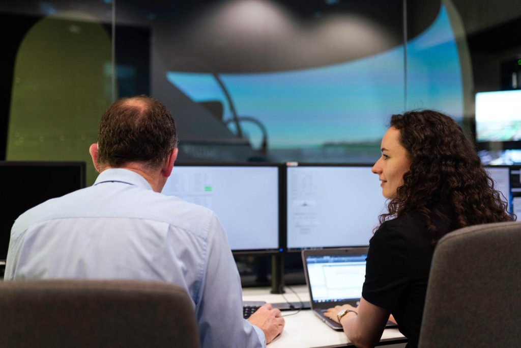 Zwei Mitarbeiter unterhalten sich in einem Kontrollturmraum mit einem riesigen Touchscreen-Display im Hintergrund